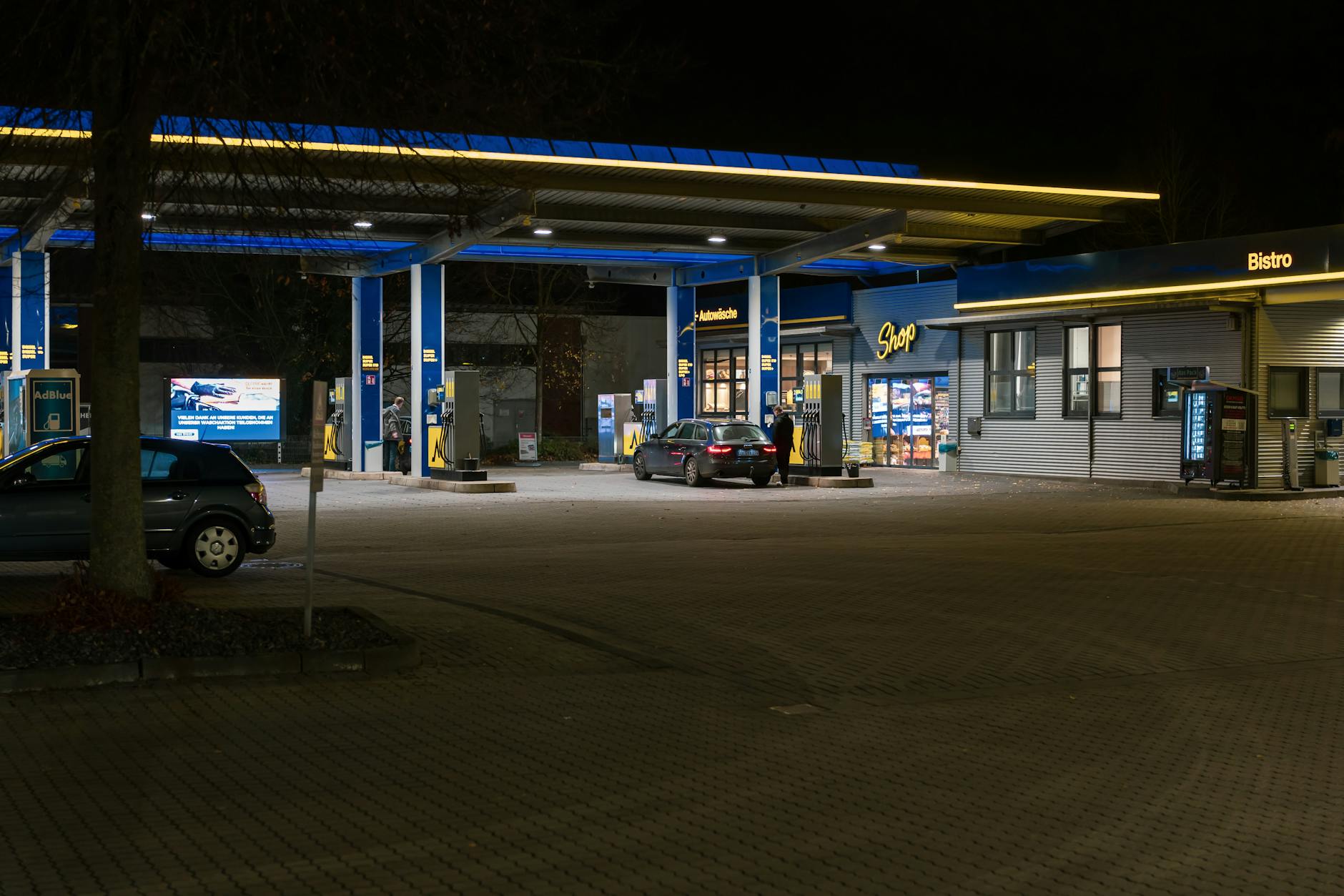 A well-lit gas station at night featuring cars and a bistro, creating a lively urban scene.