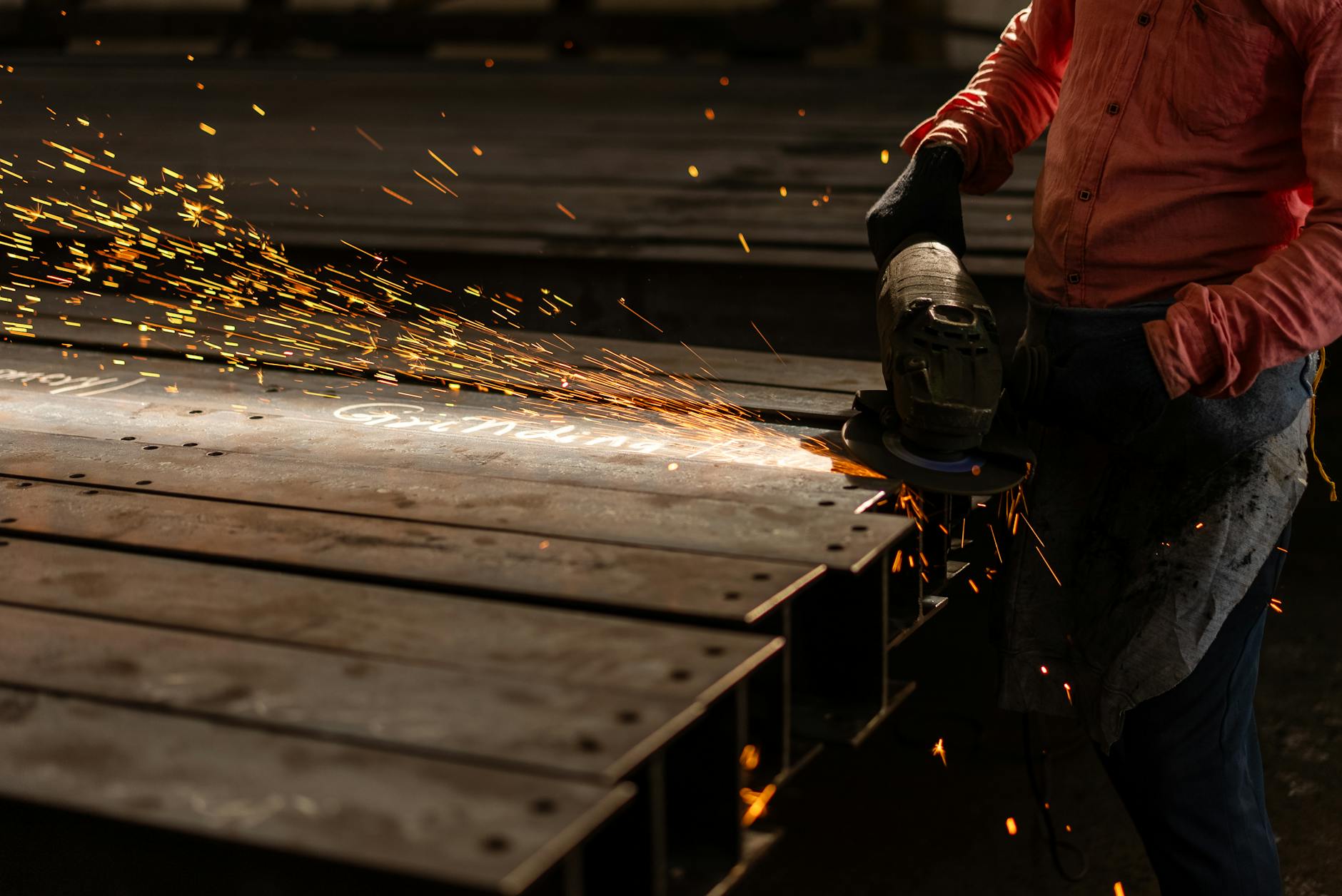 A worker grinds metal beams with an angle grinder, creating vibrant sparks in an industrial setting.