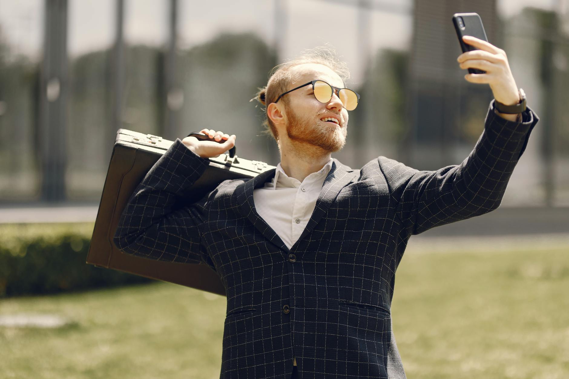 Young bearded man in a suit taking a selfie with his smartphone, holding a briefcase outdoors.