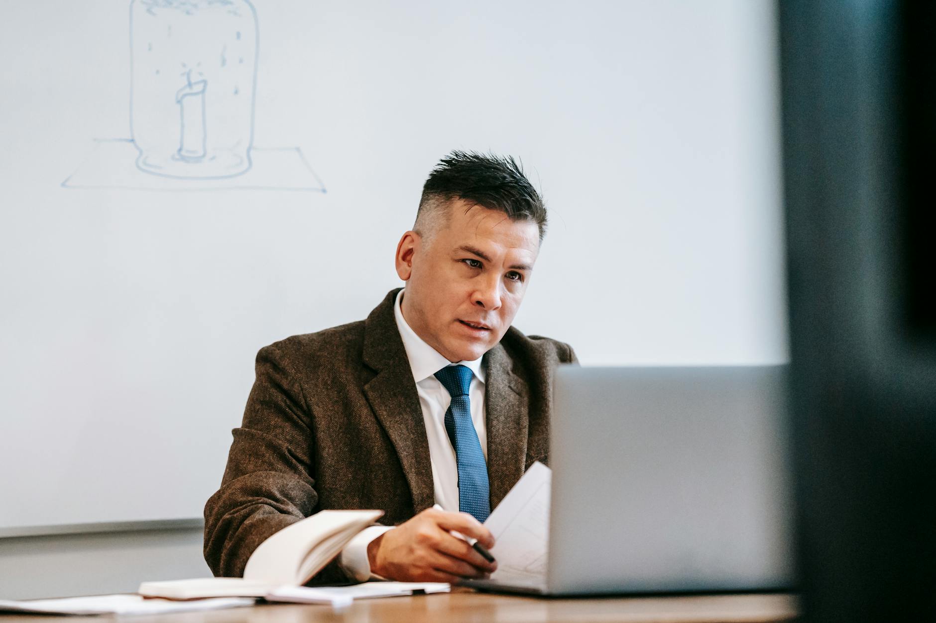 A focused professional man in a corporate setting working on a laptop with paperwork.