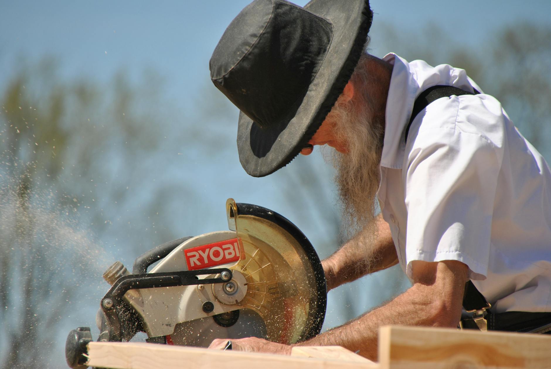 An elderly carpenter in a black hat uses a circular saw outdoors, generating sawdust.