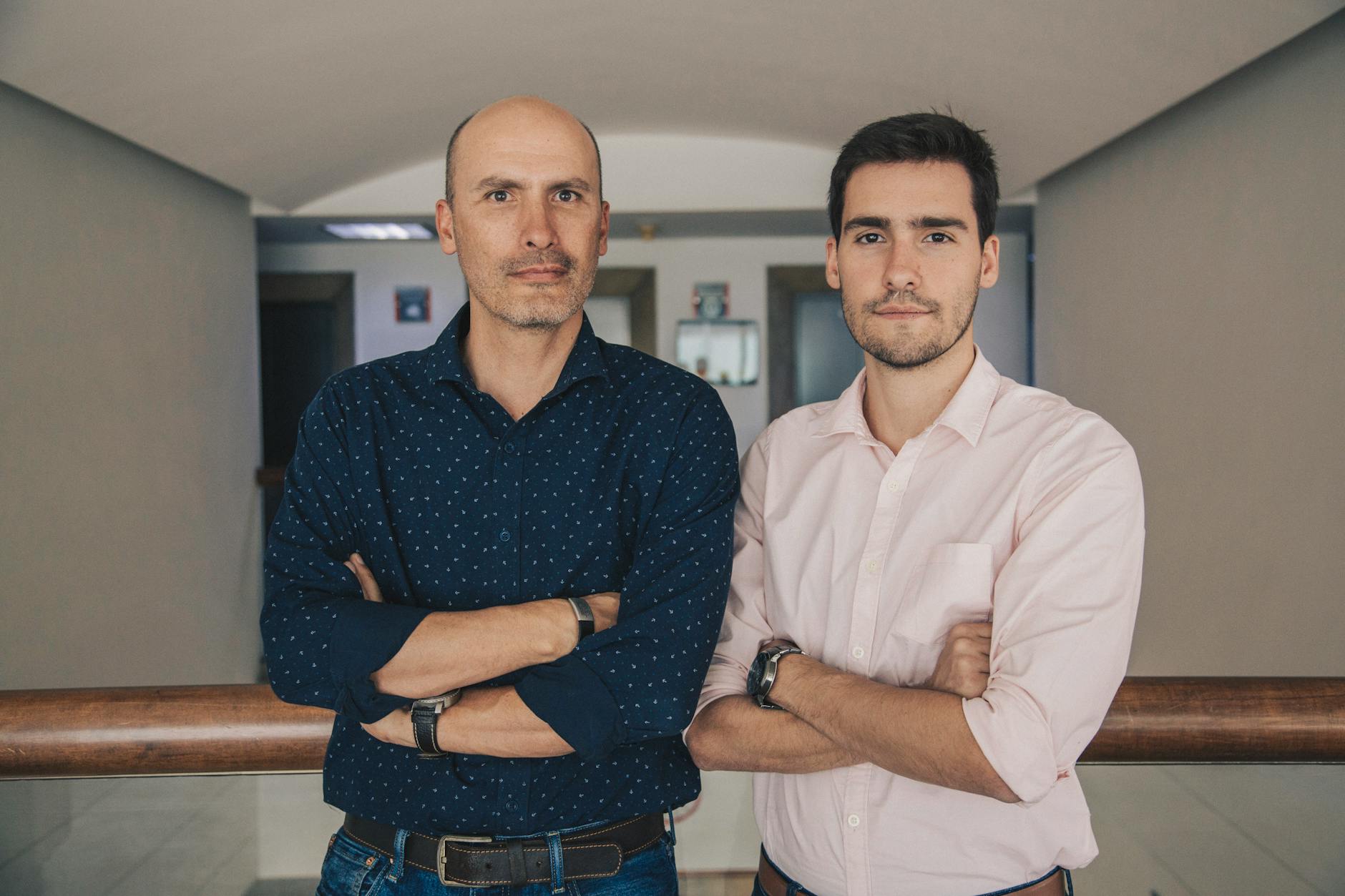Calm male colleagues in stylish shirts with wristwatches standing together with crossed arms in light modern corridor while looking away