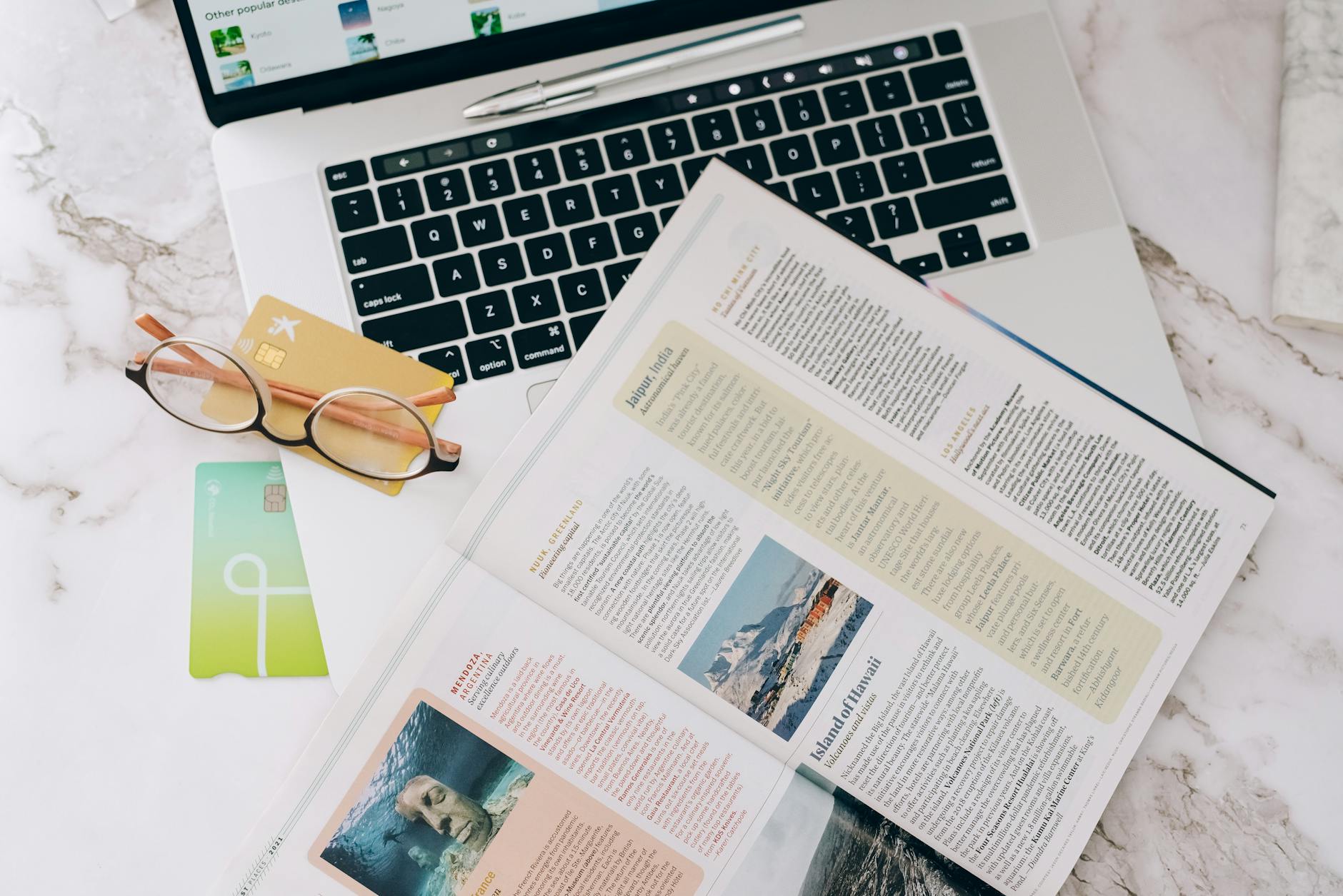 Flat lay image featuring a laptop, open magazine, credit cards, and eyeglasses on a marble table.