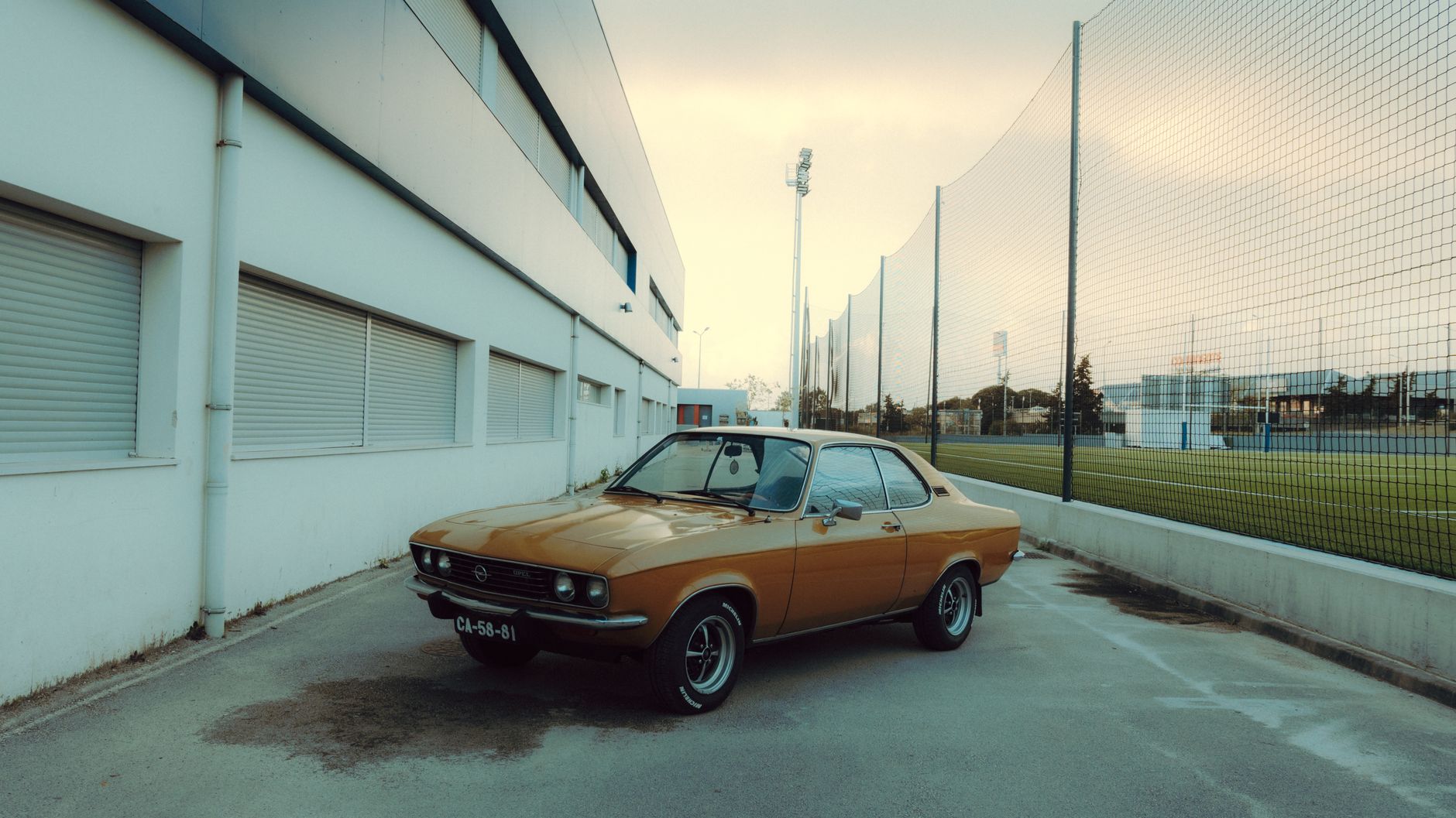 Classic orange Opel car parked outside a sports complex on a sunny day.